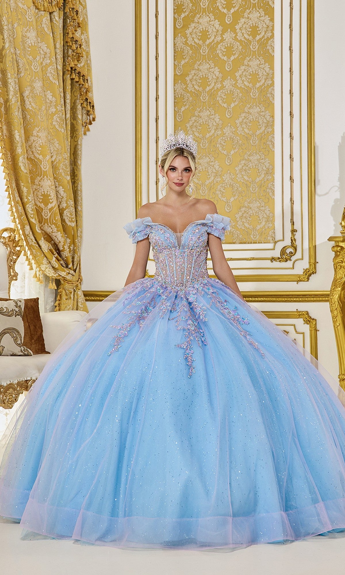 A woman stands in an ornate room wearing the Quinceanera Dress 5084 by Dancing Queen in lilac/Bahama blue with sparkling embellishments and a tiara.